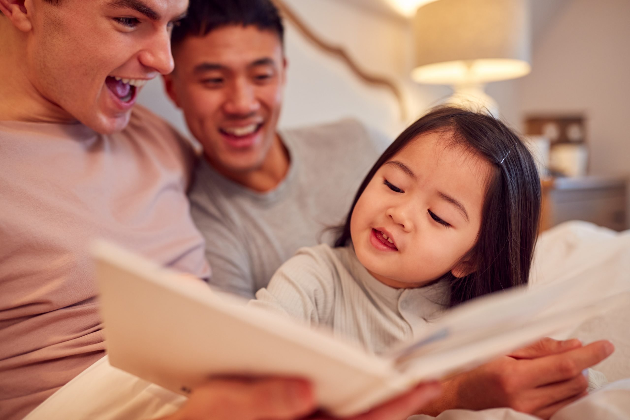 family with two dads in bed at home reading story to daughter